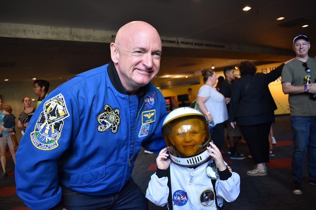 Mark Kelly, wearing his NASA flight jacket, poses for a picture with a young kid wearing an astronaut costume and space helmet.